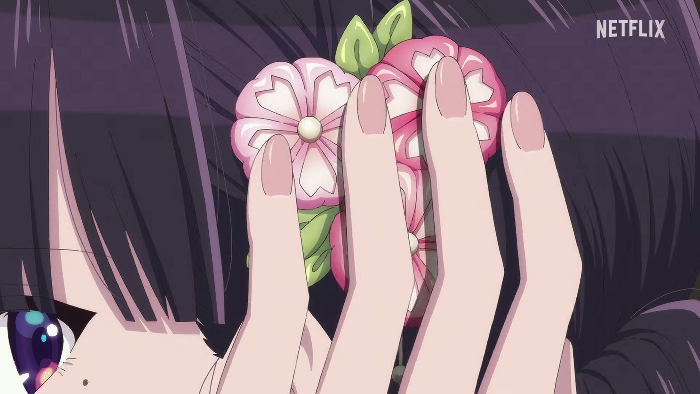A hand holding a pink and white fabric flower kanzashi with pearl centres, with Miyo's eye and beauty mark visible at the lower left of the frame