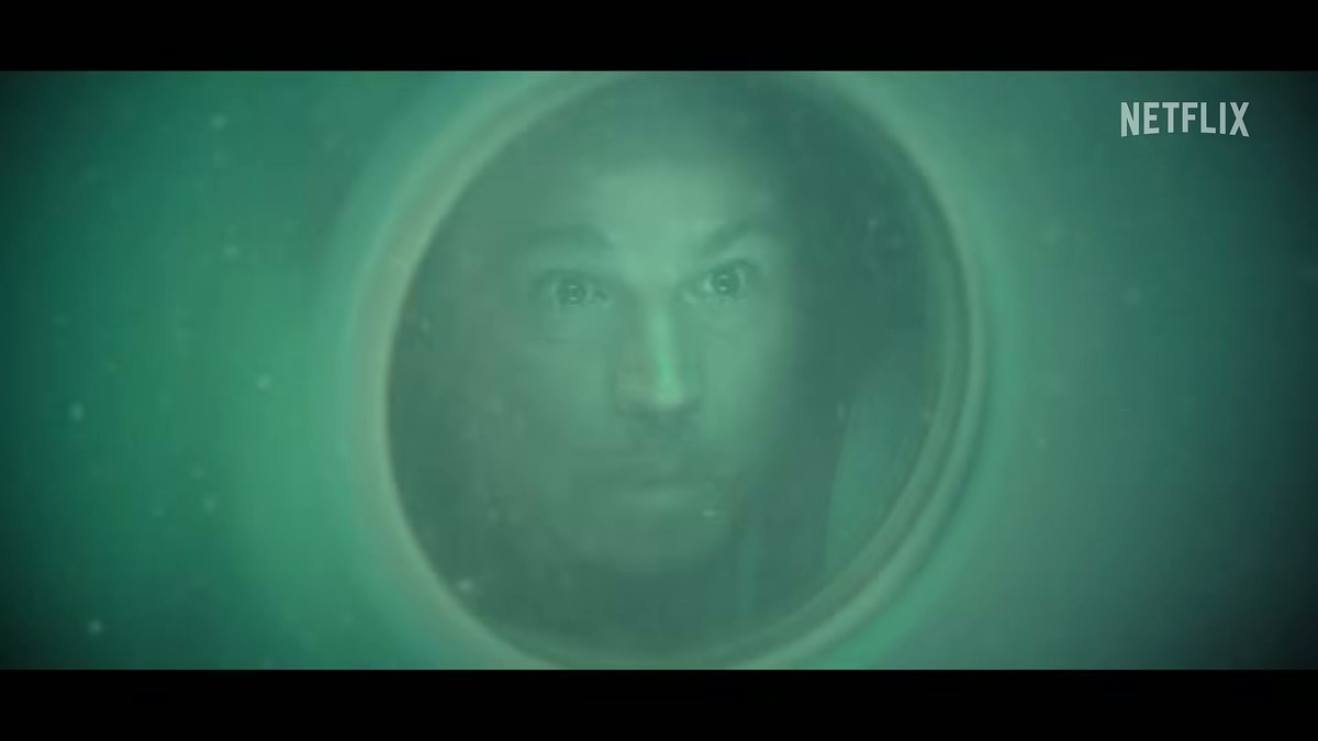 A human face lit green behind a round porthole in the Below trailer